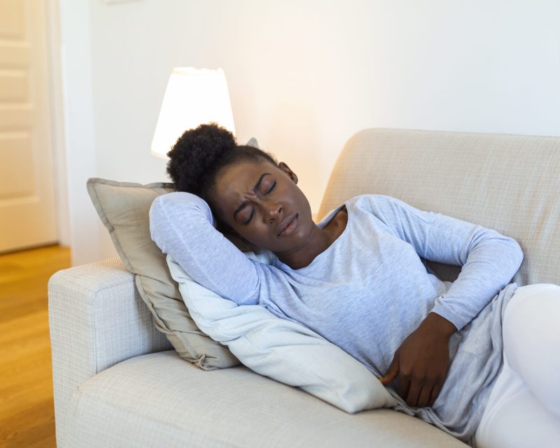 Woman lying on sofa looking sick in the living room. Beautiful young woman lying on bed and holding hands on her stomach. Woman having painful stomachache on bed, Menstrual period